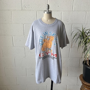May include: A gray t-shirt with a graphic of a muscular man holding a beer. The text on the shirt reads "BREWSKYS GYM" and "BUILDING BODIES TWELVE OUNCES AT A TIME". The shirt is displayed on a mannequin.