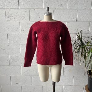 May include: A long-sleeved sweater in a deep, rich red hue. The garment features a wide, boat-style neckline and a textured, fuzzy knit construction. The sweater is displayed on a neutral-toned mannequin, showcasing its straight hem and sleeves.