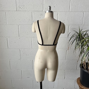 May include: A black bralette with thin straps and a low back is displayed on a mannequin. The bralette is made of a soft, dark fabric. The mannequin is set against a white brick wall with a potted plant in the background.