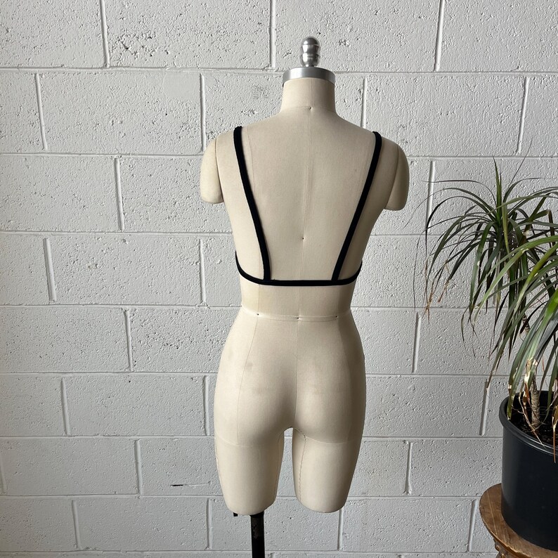 May include: A black bralette with thin straps and a low back is displayed on a mannequin. The bralette is made of a soft, dark fabric. The mannequin is set against a white brick wall with a potted plant in the background.