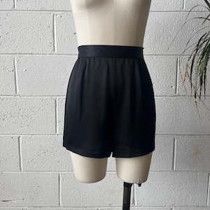 May include: Black high-waisted shorts with a wide waistband. The shorts are made of a smooth, possibly satin-like fabric. The garment is displayed on a mannequin against a white brick wall.