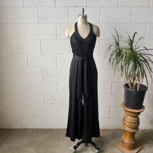 May include: A solid black sleeveless jumpsuit featuring a halter neckline with gathered fabric at the bust, a fitted waist with a matching fabric tie belt, and wide-leg trousers. The garment is displayed on a mannequin against a white brick wall.