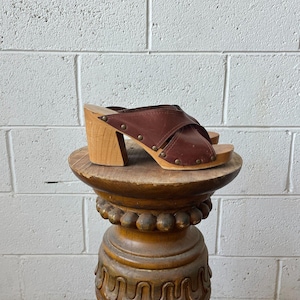 May include: A pair of brown leather clogs featuring a crisscross strap design, a light-colored wooden block heel, and a wooden sole. The leather upper is secured to the wooden base with small, evenly spaced metal studs along the sides.