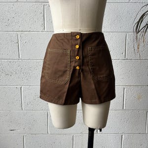 May include: Brown shorts with two front pockets and a button-up closure. The shorts have yellow stitching and are displayed on a mannequin. The shorts are a vintage style.