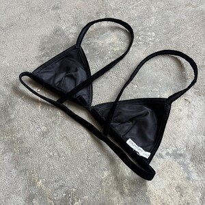May include: A black triangle-shaped bra with thin straps. The bra is made of a soft, velvety material and features a tag with the text "courreges oo". The bra is designed with a minimalist aesthetic.