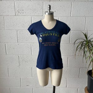 May include: A navy blue, short-sleeved t-shirt with a v-neck. The shirt features the text "I'm Perfectly Adjusted, so don't touch the knobs" in a colourful, playful font. The shirt is displayed on a mannequin.