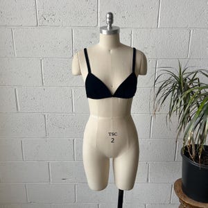 May include: A black velvet triangle bra with thin straps is displayed on a beige dress form. The dress form has the text "TSC 2" printed on the front. The background is a white brick wall and a potted plant.