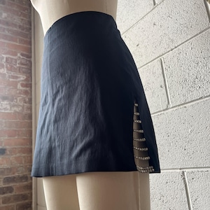 May include: A black mini skirt with a high waist and a side slit. The slit is embellished with a vertical line of sparkling rhinestones. The skirt is displayed on a mannequin.