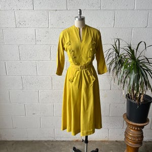 May include: A mustard yellow midi dress featuring three-quarter length sleeves, a V-neckline, and a button-down bodice detail. The dress includes a matching fabric belt with a rectangular buckle at the waist and a flared skirt silhouette.