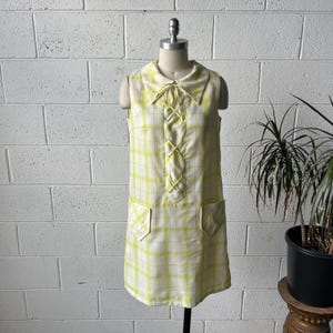 Könnte beinhalten: Ein ärmelloses Vintage-Kleid in einem gelb-weiß karierten Muster. Das Kleid hat einen Kragen mit Bindedetail, zwei Vordertaschen und einen knielangen Saum. Das Kleid wird auf einer Schaufensterpuppe präsentiert.