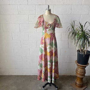 May include: A floor-length dress featuring a patchwork pattern of various floral and paisley prints in shades of pink, green, yellow, and white. The garment has short flutter sleeves, a square neckline, and a gathered bodice with a central fabric rose.