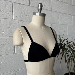 May include: A black velvet bra with thin straps is displayed on a mannequin. The bra has a triangle cup design and a simple band. The background is a white brick wall with a plant in the corner.