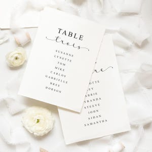 Peut inclure: Deux cartons de table ivoire avec texte noir. La première carte indique "TABLE trois" avec une liste de noms en dessous. La deuxième carte a une liste de noms. Fleurs et rubans blancs en arrière-plan.