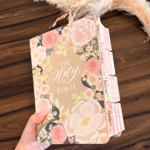 May include: A Holy Bible with a tan cover featuring a floral pattern of pink, white, and yellow flowers. The text The Holy Bible is printed in white on the front cover. The book includes multiple tabbed dividers along the side edge.