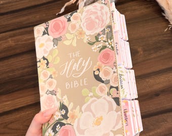May include: A Holy Bible with a tan cover featuring a floral pattern of pink, white, and yellow flowers. The text The Holy Bible is printed in white on the front cover. The book includes multiple tabbed dividers along the side edge.