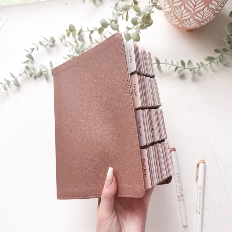 Puede incluir: Una Biblia de color rosa polvoriento con pesta&ntilde;as etiquetadas. Las pesta&ntilde;as son de diferentes tonos de rosa y tienen texto. Dos bol&iacute;grafos blancos con detalles dorados son visibles en el fondo. La Biblia est&aacute; abierta, revelando las p&aacute;ginas interiores.