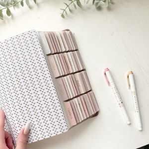 Puede incluir: Cuaderno abierto con un patr&oacute;n de tri&aacute;ngulos dorados y pesta&ntilde;as en tonos rosa y beige. Dos resaltadores blancos con tapas rosas y beige est&aacute;n junto al cuaderno. Las pesta&ntilde;as est&aacute;n etiquetadas con texto dorado.