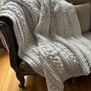 May include: A white crocheted throw blanket with a cable knit pattern draped over a wooden chair.