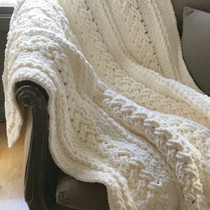 May include: A cream-colored, chunky knit blanket draped over a dark brown armchair. The blanket features a thick, textured cable knit pattern. The soft, plush material creates a cozy and inviting aesthetic. The blanket is a home decor item.