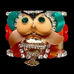 Puede incluir: Un brazalete de color con forma de búho con pedrería y detalles de gemas verdes. El búho está hecho de metal dorado y tiene un diseño detallado.