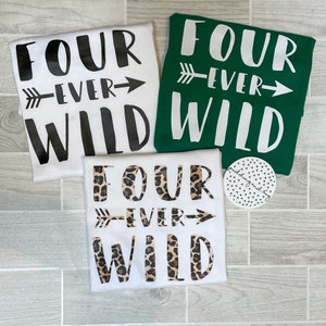 Könnte beinhalten: Drei weiße T-Shirts mit dem Text "Four Ever Wild" in Schwarz, Weiß und Leopardenmuster. Die Shirts liegen auf einem Holzgrund.