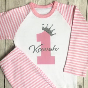 Könnte beinhalten: Rosa-weiß gestreiftes Langarm-Shirt und Hosen-Set mit einer pinkfarbenen Zahl eins und einer silbernen Glitzerkrone. Der Name "Keevah" steht unter der Zahl eins.