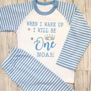 Puede incluir: Pijama blanco con rayas azules y blancas. La parte superior tiene un ribete azul y dice "When I wake up I will be One NOAH" con una corona plateada y estrellas.