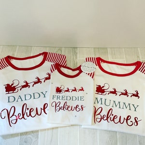 Könnte beinhalten: Drei weiße Langarmshirts mit rot-weiß gestreiften Ärmeln. Die Shirts haben einen roten Saum am Hals und ein rotes Weihnachtsmann-Motiv mit Rentieren. Der Text auf den Shirts lautet "Daddy Believes", "Freddie Believes" und "Mummy Believes".