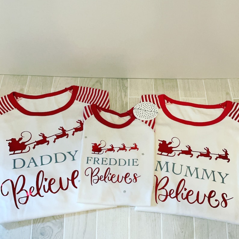 Puede incluir: Tres camisas blancas de manga larga con mangas a rayas rojas y blancas. Las camisas tienen un ribete rojo alrededor del cuello y un dise&ntilde;o rojo de Pap&aacute; Noel con renos. El texto en las camisas dice "Daddy Believes", "Freddie Believes" y "Mummy Believes".