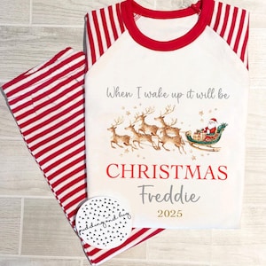 Puede incluir: Pijama de Navidad blanco con rayas rojas y blancas. La parte superior presenta un gráfico de Papá Noel en un trineo tirado por renos con el texto "When I wake up it will be Christmas Freddie 2025".