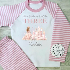 Puede incluir: Un conjunto de pijama infantil a rayas rosas y blancas. La parte superior tiene mangas rosas y un gráfico de una princesa, un castillo y el texto "When I wake up I will be THREE" y el nombre "Sophia".