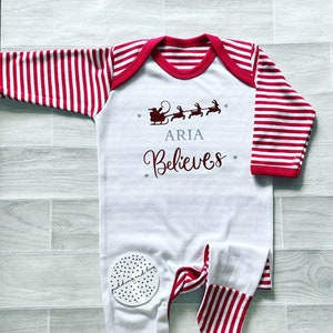 Puede incluir: Mono blanco para beb&eacute; con mangas a rayas rojas y blancas. El mono tiene un ribete a rayas rojas y blancas alrededor del cuello. El mono tiene un gr&aacute;fico de Pap&aacute; Noel en un trineo con renos y el texto "ARIA Believes".