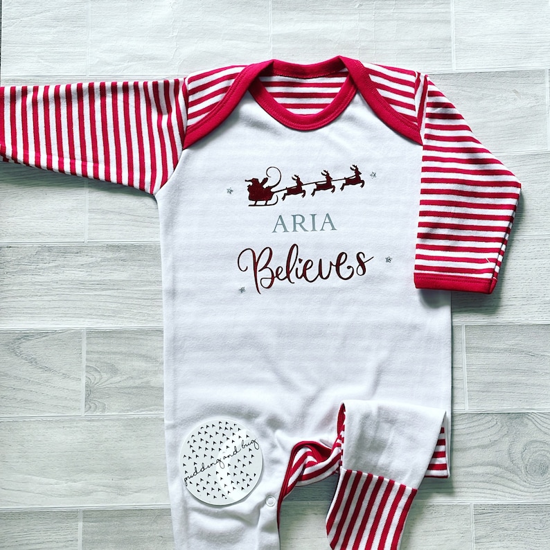 Puede incluir: Mono blanco para beb&eacute; con mangas a rayas rojas y blancas. El mono tiene un ribete a rayas rojas y blancas alrededor del cuello. El mono tiene un gr&aacute;fico de Pap&aacute; Noel en un trineo con renos y el texto "ARIA Believes".