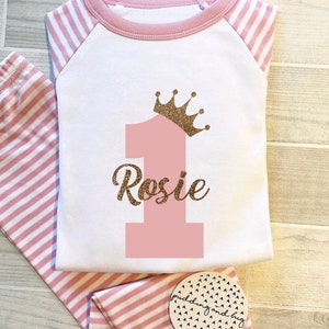 Puede incluir: Pijama a rayas rosa y blanco con un número uno rosa y una corona dorada con purpurina en la parte superior. El nombre "Rosie" está escrito en purpurina dorada debajo del número uno.