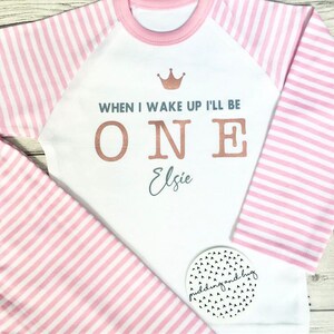 K&ouml;nnte beinhalten: Wei&szlig;er Langarm-Strampler mit rosa-wei&szlig;en Streifen an den &Auml;rmeln und einer rosa Krone auf der Vorderseite. Der Strampler sagt "When I wake up I'll be ONE Elsie" in goldenen und silbernen Buchstaben.