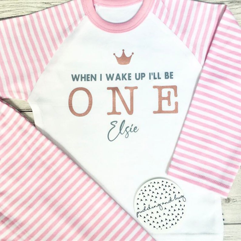 K&ouml;nnte beinhalten: Wei&szlig;er Langarm-Strampler mit rosa-wei&szlig;en Streifen an den &Auml;rmeln und einer rosa Krone auf der Vorderseite. Der Strampler sagt "When I wake up I'll be ONE Elsie" in goldenen und silbernen Buchstaben.