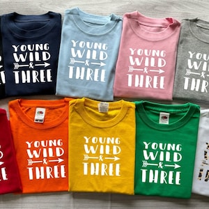 Könnte beinhalten: Zehn T-Shirts in verschiedenen Farben mit dem Aufdruck "Young Wild & Three" auf der Vorderseite. Die Shirts sind in zwei Reihen zu fünf angeordnet.