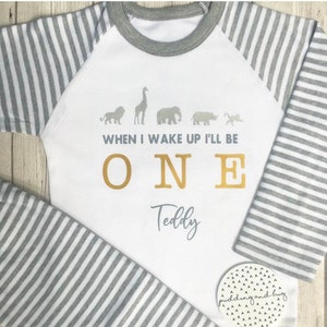 Könnte beinhalten: Weißer Baby-Strampler mit grauen Streifen an den Ärmeln. Der Strampler hat ein goldenes Tiermotiv und den Text "When I wake up I'll be ONE Teddy".