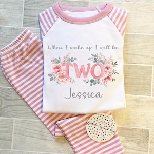 Puede incluir: Pijama a rayas rosa y blanco con una camisa blanca de manga larga que dice "Cuando me despierte seré DOS Jessica". La camisa tiene un diseño floral con flores rosas y hojas verdes.