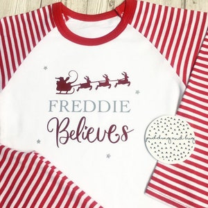 Puede incluir: Camiseta blanca de manga larga con rayas rojas y blancas en las mangas. La camiseta tiene un cuello redondo rojo y un dise&ntilde;o rojo y burdeos en la parte delantera que dice "FREDDIE Believes" con una silueta de Pap&aacute; Noel en un trineo tirado por renos.