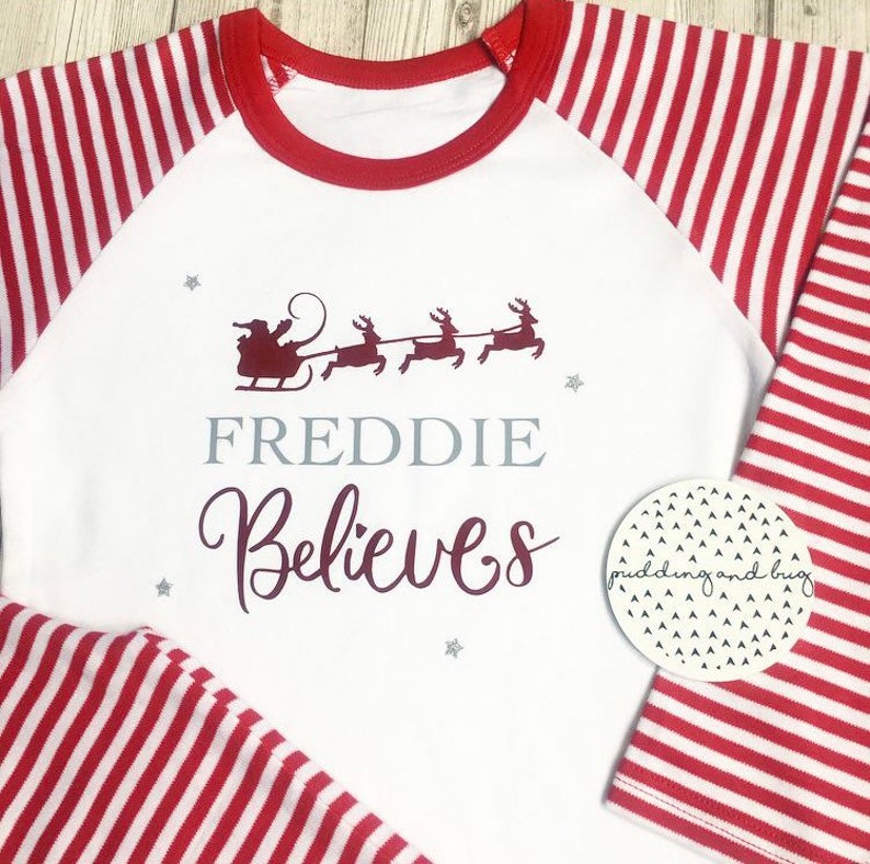 Puede incluir: Camiseta blanca de manga larga con rayas rojas y blancas en las mangas. La camiseta tiene un cuello redondo rojo y un dise&ntilde;o rojo y burdeos en la parte delantera que dice "FREDDIE Believes" con una silueta de Pap&aacute; Noel en un trineo tirado por renos.