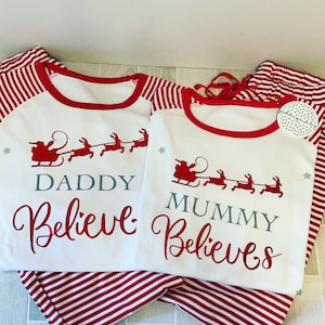Puede incluir: Pijama a rayas rojas y blancas con una camisa blanca de manga larga con un dise&ntilde;o de Pap&aacute; Noel y renos. La camisa dice "Daddy Believe" y "Mummy Believes" en texto gris.