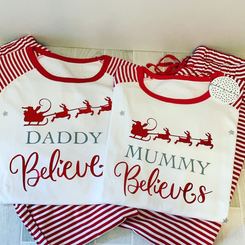 Puede incluir: Pijama a rayas rojas y blancas con una camisa blanca de manga larga con un dise&ntilde;o de Pap&aacute; Noel y renos. La camisa dice "Daddy Believe" y "Mummy Believes" en texto gris.
