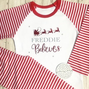 Puede incluir: Pijama a rayas blancas y rojas con un ribete rojo alrededor del cuello. La parte delantera de la camisa tiene una ilustraci&oacute;n roja de Santa Claus en su trineo con renos. El texto "FREDDIE Believes" est&aacute; impreso en gris y burdeos en la camisa.