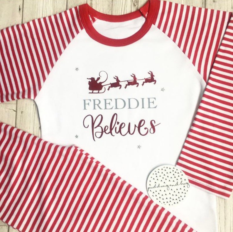 Puede incluir: Pijama a rayas blancas y rojas con un ribete rojo alrededor del cuello. La parte delantera de la camisa tiene una ilustraci&oacute;n roja de Santa Claus en su trineo con renos. El texto "FREDDIE Believes" est&aacute; impreso en gris y burdeos en la camisa.
