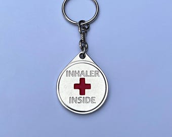 May include: A silver keyring with a round tag that says "INHALER INSIDE" with a red cross in the centre.