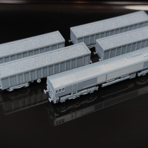 Puede incluir: Un conjunto de cuatro vagones de tren a escala de color gris sin pintar, que incluye una locomotora y tres vagones de carga abiertos. Cada vagón presenta una carga texturizada que se asemeja a la grava, mientras que la locomotora muestra un cuerpo largo y rectangular con bogies de ruedas detallados y mecanismos de acoplamiento en ambos extremos.