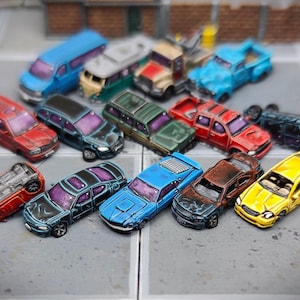 May include: A collection of 13 miniature toy cars in various colours, including blue, red, yellow, green, and black. The cars are in various states of damage, with some overturned and others with missing parts. The cars are arranged on a grey and white chequered surface.