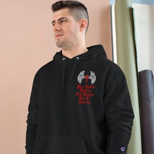 May include: Black hooded sweatshirt with a front pocket and a Champion logo on the left sleeve. The sweatshirt features a red cross with angel wings and text that reads "Deus Meus Dominus, Sed Humilis, Sed Et Gloriosus."