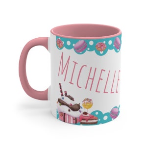 May include: White ceramic mug with a pink rim and handle. The mug has a blue and white design with a variety of sweets and the name "Michelle" written in pink.